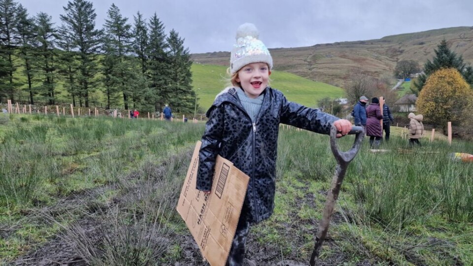 young child with a  big smile and a bobble hat on holding a spade after planting a tree